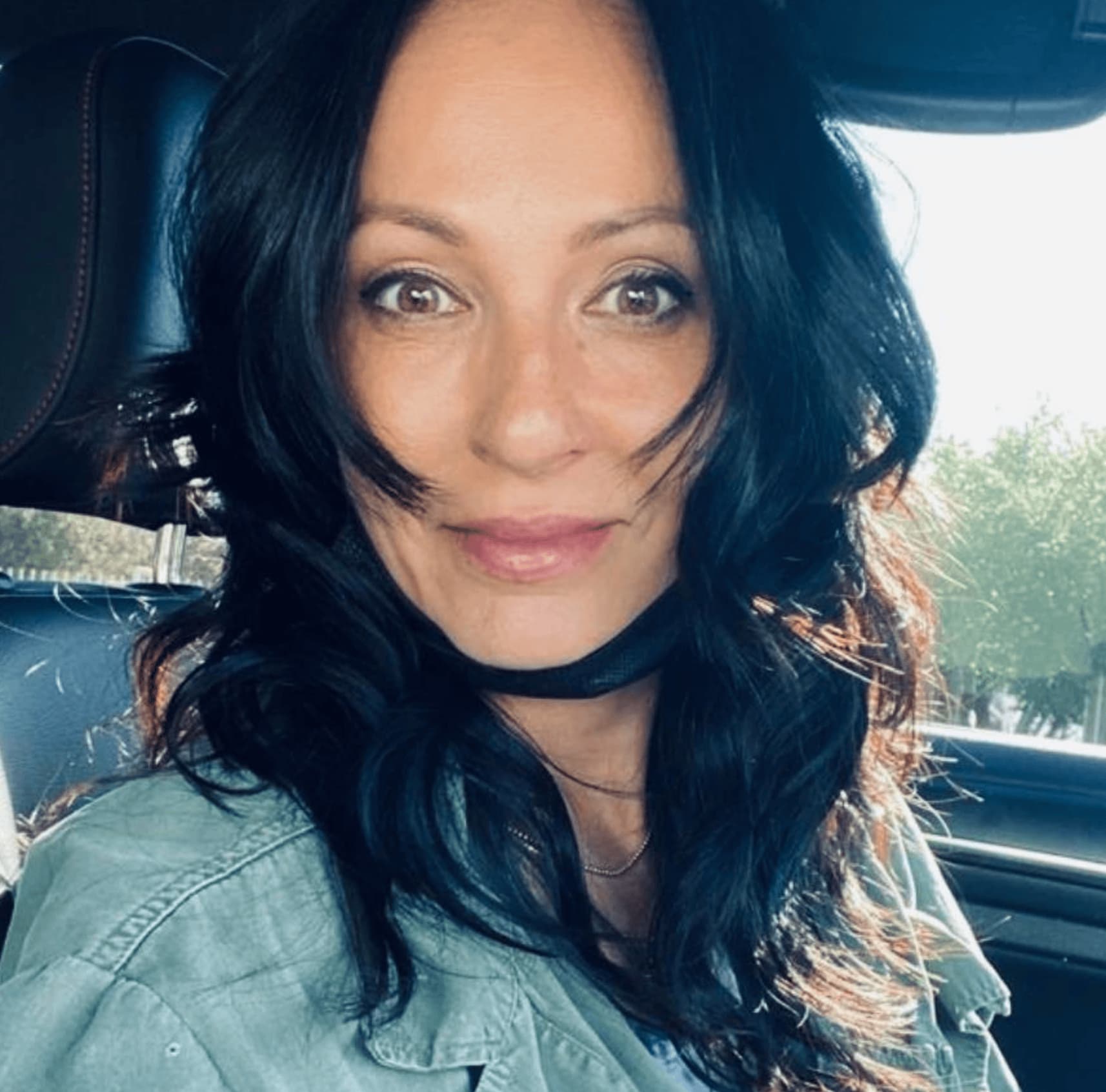 Woman with long dark hair sitting in a car, wearing a green jacket, smiling, and looking at the camera.