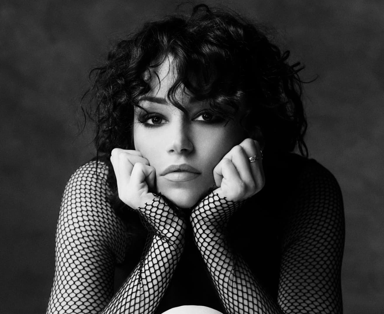 Black and white photo of person with curly hair resting chin on hands, wearing fishnet sleeves.